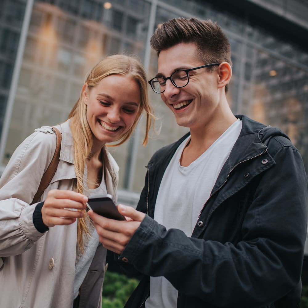 A young man and woman are standing outside and laughing while looking at a phone.