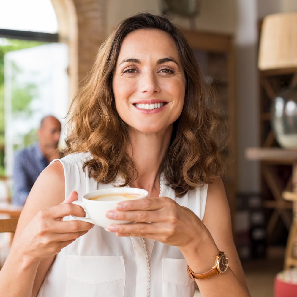 A woman smiles showing off her white teeth while holding a cup of coffee.